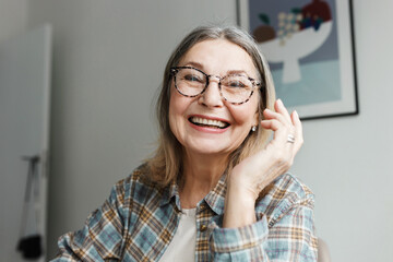 Portrait of happy laughing grandmother in glasses and plaid shirt video chatting with her grandchild online, sitting at home in living-room, having fun, enjoying nice and warm conversation