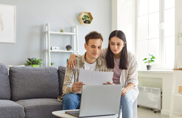 Portrait of young couple sitting on sofa with laptop looking through financial documents at home....