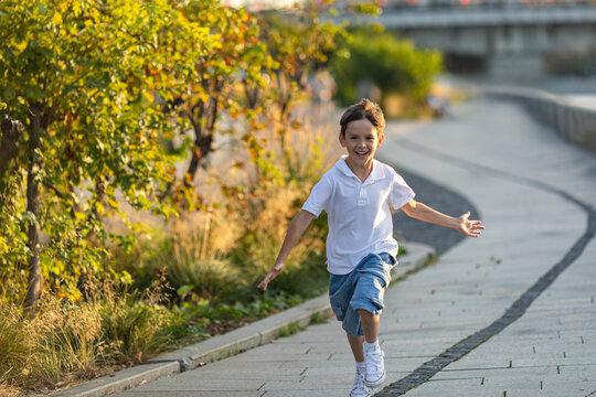 A happy 10 year old boy with outstretched arms runs on the asphalt on the city embankment along the river. Carefree childhood, freedom and summer holidays concept.