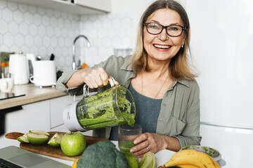 Happy cheerful aged female pouring green detox smoothie in glass, having detox nutritious breakfast as antioxidant support to stay healthy and prolong beauty, . Vegetarian diet and weight loss program