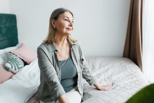 Happy adorable retired lady in casual clothes admiring cityscape sitting on bed and looking through window in day time, enjoying retirement, relaxing and having rest after fulfilling domestic chores
