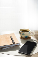 A workspace featuring a cup of coffee, a smartphone on stacked notebooks, a black pen, and a blurred green plant on a wooden desk. Natural lighting and shadows create a cozy and productive atmosphere.