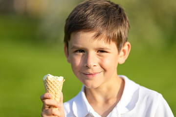 A happy smiling child boy in white t-shirt eats ice cream on a hot summer day in park on green...