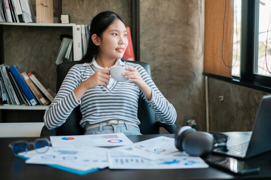 Young woman in striped shirt taking a coffee break in a modern office setting with documents and a laptop.