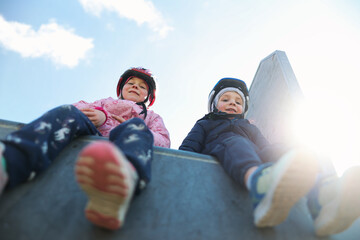 A boy and a girl sit on the ramp at a skate park, taking a break and enjoying the lively atmosphere around them.