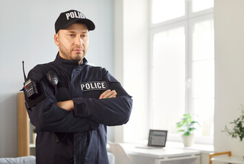 Portrait of confident male police officer standing in office wearing uniform with walkie talkie with copy space. Young policeman with crossed arms standing indoors. Crime and robbery concept.