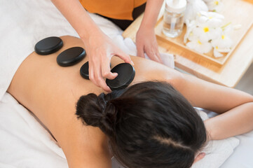 Hot stone massage therapy concept. Therapist in orange placing black basalt stones on woman back at spa salon