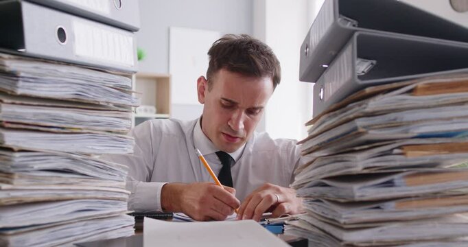 Overworked business man sitting at the desk on workplace and looking at a pile of folders and documents. Portrait of tired company employee accountant working in office. 4k video. Slow motion video.