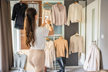 A young woman photographs clothing hanging on a wardrobe in bedroom using her smartphone. Online resale and second-hand fashion thrive through mobile apps, making sustainable shopping more accessible