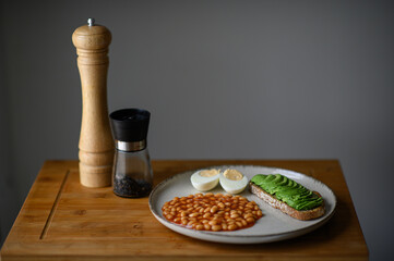 A homemade breakfast featuring avocado toast on whole grain bread, baked beans in tomato sauce, and a boiled egg. A salt shaker and pepper mill sit nearby, completing the cozy and nutritious meal.
