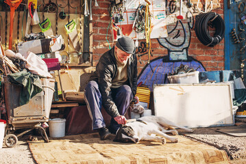 An older man dressed in old ragged clothes combs a dog at a flea market among the trash and other...