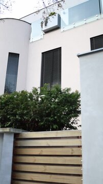 Vertical screen: Modern residential building with rounded corner, vertical windows, balcony glass panels and greenery behind a wooden fence in vertical format