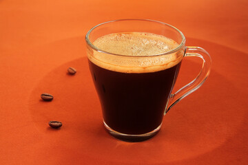 Concept, hot, aromatic, black coffee with thick foam in a glass cup on a yellow tablecloth. Coffee beans are scattered around the cup, symbolizing the time of drinking the invigorating drink.