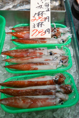 Fresh squid neatly arranged in green baskets on crushed ice at a Tokyo fish market, with handwritten price tags indicating their availability for sale