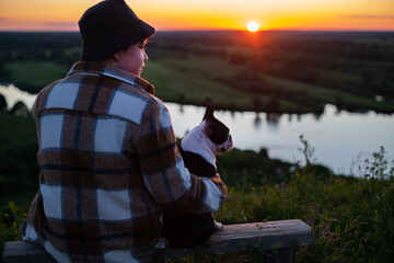 A boy sits on a bench and communicates with his friend, a Boston Terrier dog, in nature at sunset in summer.