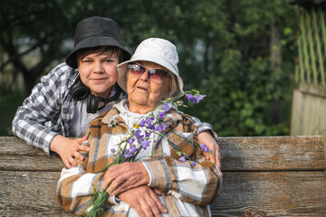 Happy grandson hugs old great-grandmother in fashionable clothes. Boy gives flowers to...