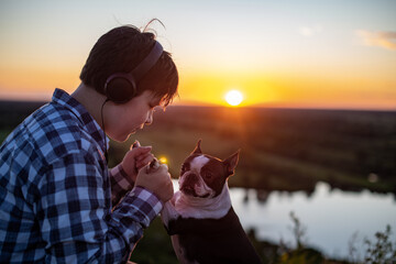 A boy sits on a bench and communicates with his friend, a Boston Terrier dog, in nature at sunset in summer.