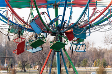 An old, non-functioning Swing Ride carousel in a public park, metal colorful chairs and chains are...