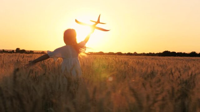 Adorable female kid silhouette running with aircraft toy at cinematic sunset sunrise wheat field back view. Happy little girl child playing pilot game with plane plaything enjoy childhood at dusk