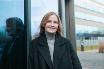 Blonde business lady girl In a stylish black coat standing near business center and looking at camera smiling. High quality photo
