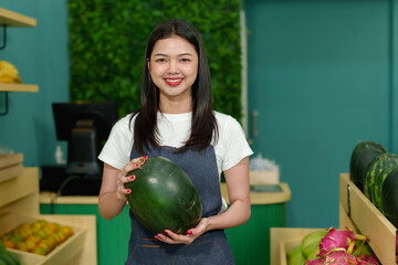 Fototapeta premium Beautiful Asian small business fruit shop owner smiling with confidence while holding green watermelon in hands wearing denim apron preparing tropical fruit shelf in fresh grocery store