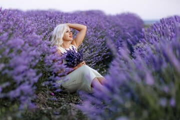 Blonde woman poses in lavender field at sunset. Happy woman in white dress holds lavender bouquet....