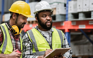 Two diverse professional male workers holding board, checking shipping stocks in storage, warehouse or factory for delivery, wearing safety hat. Diversity, Commercial Industry Business Concept.