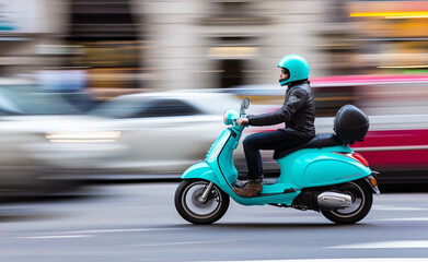 A person riding a teal scooter through a blurred urban street, captured in motion with a sense of speed.
