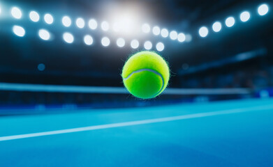 A tennis ball suspended in mid-air above a blue court during a night match under stadium lights.
