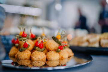 Golden breaded appetizer balls are topped with mini peppers and pickles on skewers, beautifully arranged on a shiny silver platter at an event..