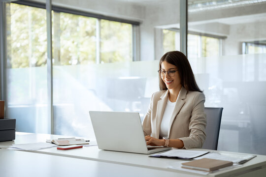 Young professional it specialist latin hispanic business lady working on laptop pc sitting at desk in modern office space. 30s middle eastern indian woman using computer technology app for work online - Powered by Adobe