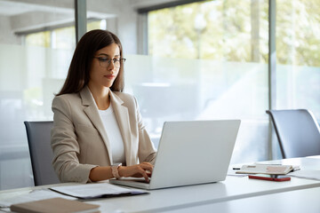 Focused financial specialist latin hispanic businesswoman working on laptop pc sitting at desk in modern office. Middle eastern or european woman using computer for finance data analysing work online