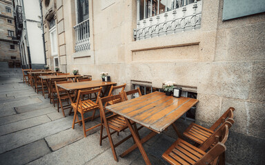 Empty outdoor cafe: wooden tables, folding chairs, flower decorations, napkin holders, stone wall background, European city street, daylight, cozy atmosphere, nobody present; Santiago de Compostela