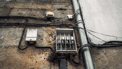 Close-up of an old electrical distribution box with open cover and exposed wiring mounted on a weathered stone wall, along with cables and conduit pipes, outdoors during daytime