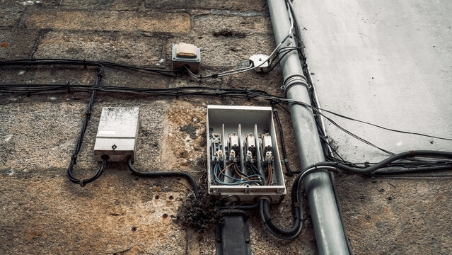 Close-up of an old electrical distribution box with open cover and exposed wiring mounted on a weathered stone wall, along with cables and conduit pipes, outdoors during daytime