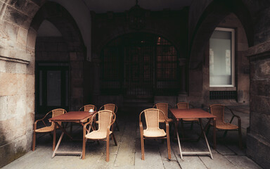 Empty outdoor cafe terrace under stone arches with wooden tables and wicker chairs, moody lighting, historic European architecture, cozy atmosphere; Santiago de Compostela, Spain