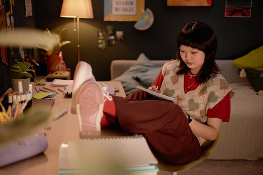 Young Asian woman sitting in cozy bedroom, focusing on her studies and relaxing on chair. Room decorated with warm lighting, books, and personal items creating a comfortable study space