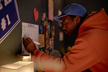 Man in orange hoodie and blue cap writing schedule on bulletin board with various items attached,...