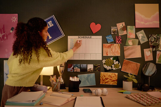 Young woman decorating desk with various pictures while organizing schedule on wall calendar on cluttered desk. Engaging in creative activity in cozy workspace