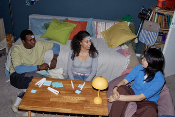Three friends enjoying playing game in cozy living room with sticky notes on their foreheads,...