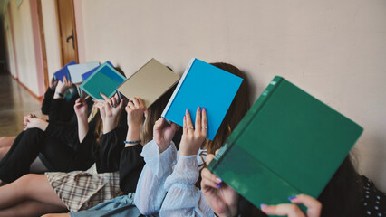 Female students sitting on the floor in a school hallway with books covering their faces