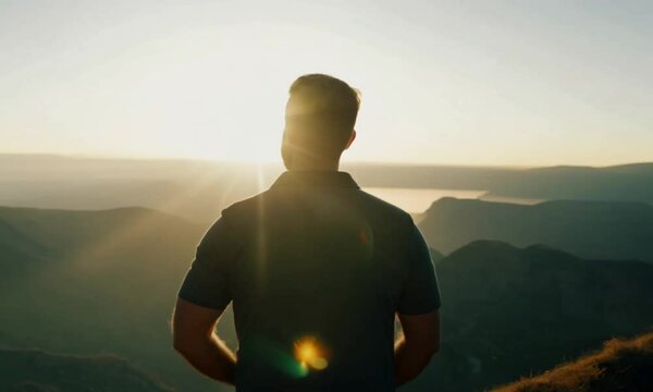 A man stands strong on a cliff&rsquo;s edge, gazing toward the horizon with determination. The sky glows with the first light of dawn. Cinematic