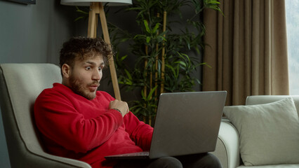 A young man in a red sweater sitting on a couch, looking surprised while using a laptop