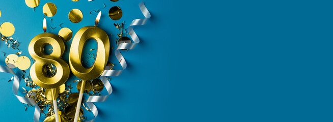 80 years celebration. Gold burning candles of number eighty on blue background with confetti, copy space.