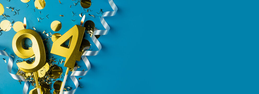 94 years celebration. Gold burning candles of number ninety-four on blue background with confetti, copy space.