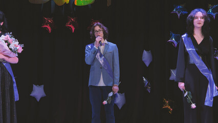 Schoolboy wearing a sash giving a speech during his graduation ceremony with his friends, holding a microphone and a rose