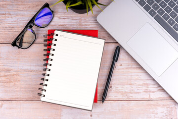 Flat lay view of a laptop, notebooks, pen, an eyeglasses and potted green plant on a wooden table