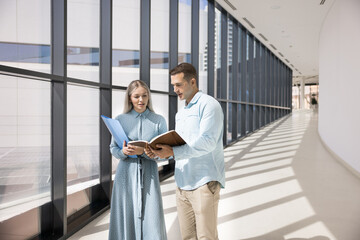 Two professionals standing in contemporary hallway looking at personal organizer records, reviewing documents, preparing for presentation, or finalizing details for joint project at break in office