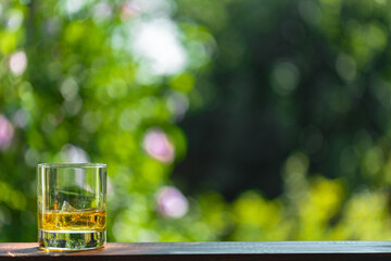 A glass of whiskyl with ice cube on a wooden table outdoors in the summer garden with space for copy.