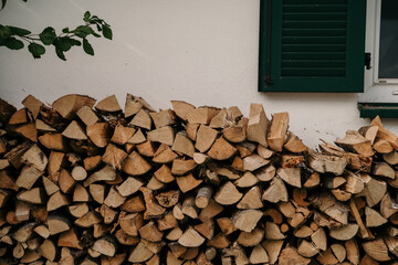irewood by the wall. Stack of firewood by the wall of a building. Visible window with green...